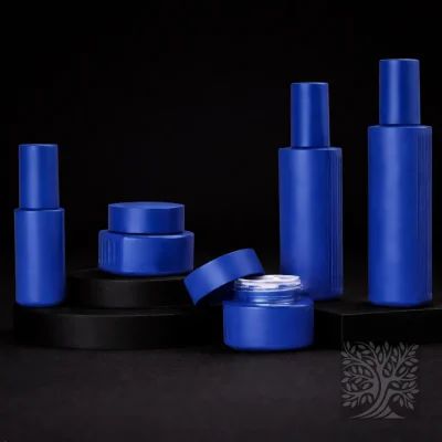 Matte cobalt blue skincare bottles and cosmetic jars with rounded forms, including an open jar, arranged on black display blocks.